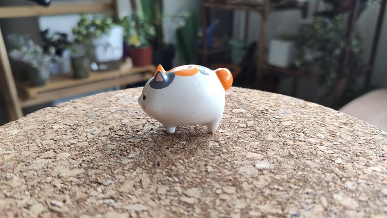 K&ouml;nnte beinhalten: Eine kleine Keramikfigur eines Katers mit wei&szlig;em Fell, orangefarbenen und schwarzen Markierungen und einem runden K&ouml;rper.