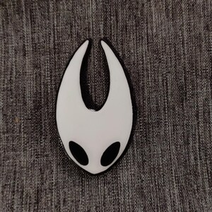 Może przedstawiać: Pin emaliowany na biało z czarnym obramowaniem w kształcie wydrążonego serca z dwoma czarnymi oczami. Pin przedstawia popularną postać z gry wideo Hollow Knight.