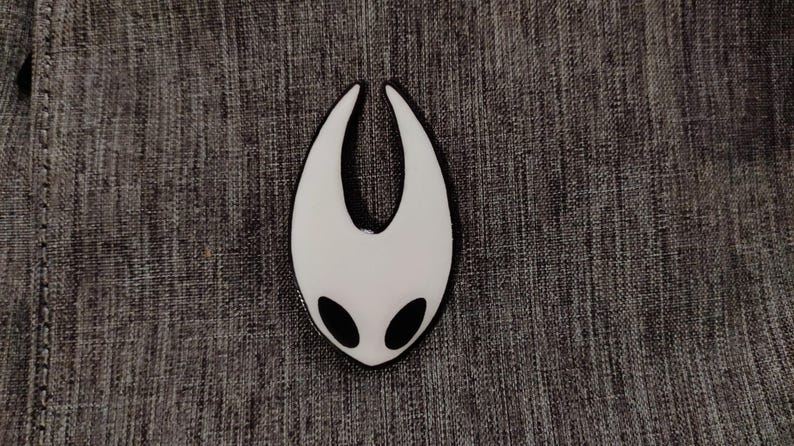 Może przedstawiać: Pin emaliowany na biało z czarnym obramowaniem w kształcie wydrążonego serca z dwoma czarnymi oczami. Pin przedstawia popularną postać z gry wideo Hollow Knight.