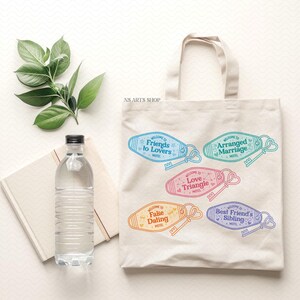 May include: A cream tote bag displaying colourful motel keychains with phrases such as "Friends to Lovers" and "Arranged Marriage." A water bottle and a notebook are next to the bag. Green leaves are at the top.