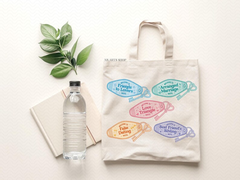 May include: A cream tote bag displaying colourful motel keychains with phrases such as "Friends to Lovers" and "Arranged Marriage." A water bottle and a notebook are next to the bag. Green leaves are at the top.