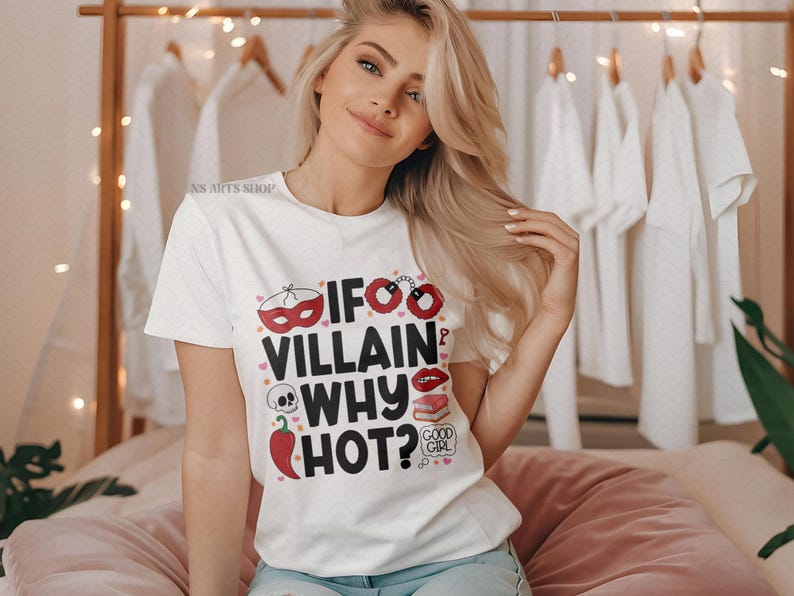 Puede incluir: Camiseta blanca con el texto "If Villain Why Hot?" en negro, rojo y rosa. La camiseta presenta elementos gr&aacute;ficos como una m&aacute;scara, esposas, labios, una calavera, un chile y un libro. La modelo lleva la camiseta.