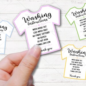 May include: A hand holding several t-shirt shaped washing instruction cards. Each card has the words "Washing Instructions" and laundry care details. The cards are outlined in blue, pink, green, and yellow.