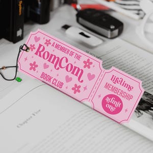 May include: A pink bookmark with the text "A MEMBER OF THE RomCom BOOK CLUB" and "Lifetime MEMBERSHIP admit one." The bookmark is decorated with hearts and flowers. A black and gold bookmark is attached to the pink bookmark.