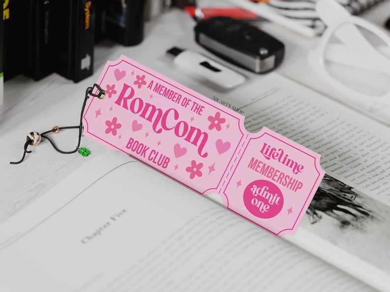May include: A pink bookmark with the text "A MEMBER OF THE RomCom BOOK CLUB" and "Lifetime MEMBERSHIP admit one." The bookmark is decorated with hearts and flowers. A black and gold bookmark is attached to the pink bookmark.