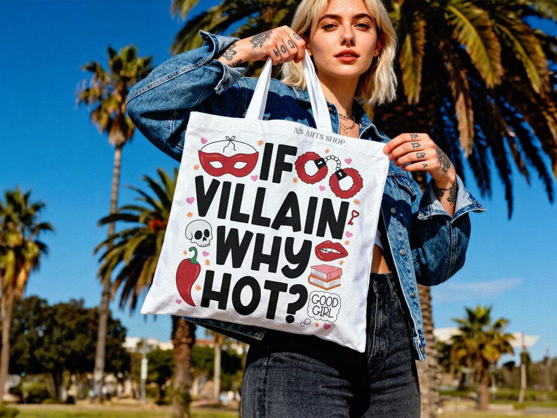 Puede incluir: Bolso tote blanco con el texto "If Villain Why Hot?" y varias ilustraciones, incluyendo una m&aacute;scara, esposas, una calavera, un chile y labios. El bolso es sostenido por una persona con una chaqueta vaquera.