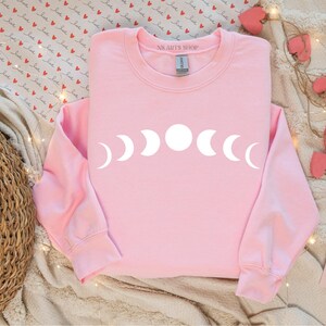 May include: Pink sweatshirt with a white moon phase design. The text "AS.ART'S SHOP" is on the neckline.