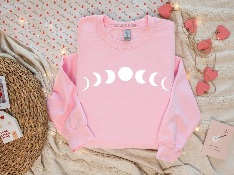 May include: Pink sweatshirt with a white moon phase design. The text "AS.ART'S SHOP" is on the neckline.