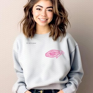 May include: A pale grey sweatshirt with long sleeves. The sweatshirt features the text "NS ARTS SHOP" on the upper left chest. A pink graphic of a motel key tag with the words "Welcome to Reverse Harem Hotel" is printed on the front.