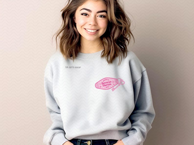 May include: A pale grey sweatshirt with long sleeves. The sweatshirt features the text "NS ARTS SHOP" on the upper left chest. A pink graphic of a motel key tag with the words "Welcome to Reverse Harem Hotel" is printed on the front.