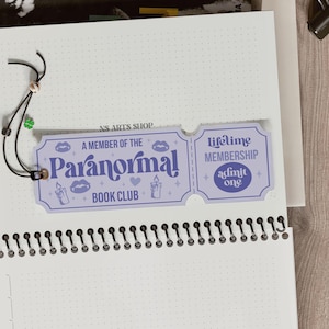 May include: A light purple "Paranormal Book Club" membership card with the text "A Member of the Paranormal Book Club" and "Lifetime Membership". The card is attached to a notebook with a black string and a green clover charm.