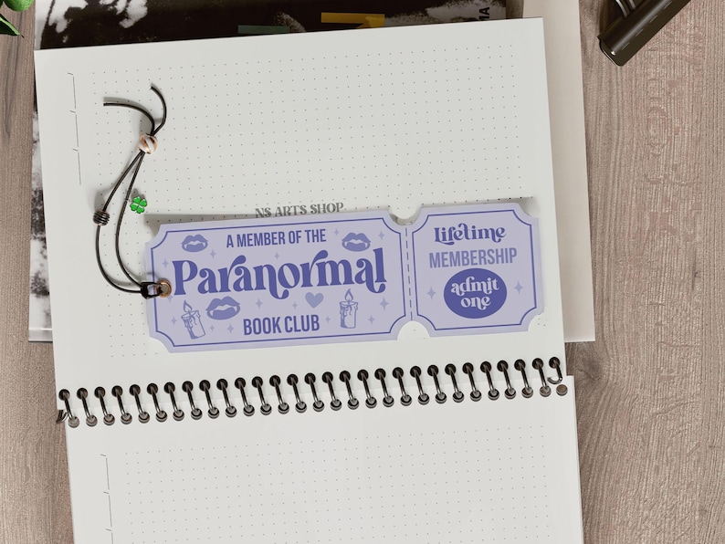 May include: A light purple "Paranormal Book Club" membership card with the text "A Member of the Paranormal Book Club" and "Lifetime Membership". The card is attached to a notebook with a black string and a green clover charm.