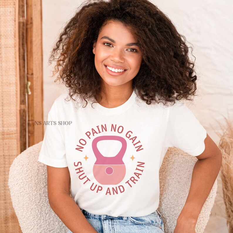 Puede incluir: Camiseta blanca con un gr&aacute;fico de pesa rusa rosa y blanca y el texto "No pain no gain shut up and train".