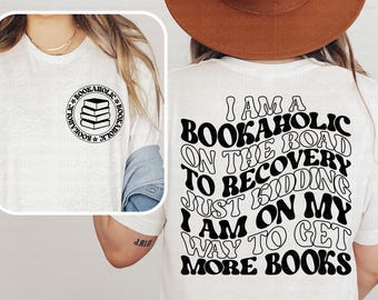 May include: A white t-shirt featuring a small circular graphic on the front left chest with a stack of books and the text BOOKAHOLIC. The back displays large black wavy text reading I AM A BOOKAHOLIC ON THE ROAD TO RECOVERY JUST KIDDING I AM ON MY WAY TO GET MORE BOOKS.