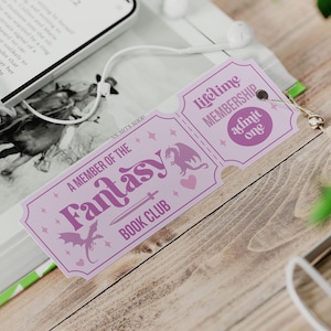 May include: A lavender-colored bookmark with the text "A MEMBER OF THE Fantasy BOOK CLUB" and "Lifetime MEMBERSHIP admit one." The bookmark features a dragon, sword, and heart design. A tassel and earbuds are also visible.