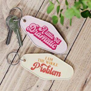 May include: Two motel-style keychains with silver key rings. One keychain is white with the text "A little bit Dramatic" in pink and red. The other is cream-colored with the text "I AM THE WHOLE PROBLEM" in red and orange.