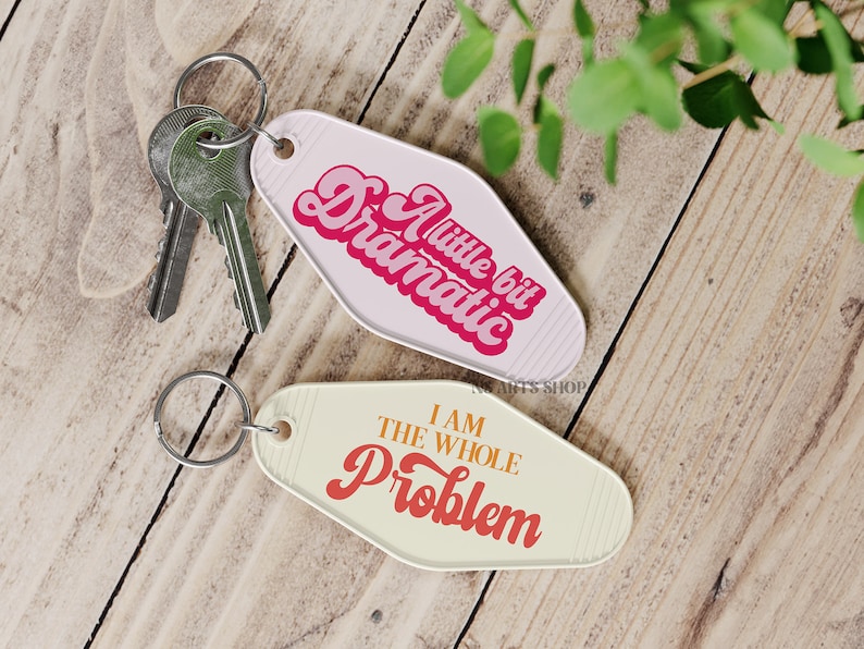 May include: Two motel-style keychains with silver key rings. One keychain is white with the text "A little bit Dramatic" in pink and red. The other is cream-colored with the text "I AM THE WHOLE PROBLEM" in red and orange.