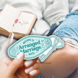 May include: A teal and white motel key tag with the words "WELCOME TO Arranged Marriage MOTEL" and a key design. The key tag is held in a hand with white nail polish. A book and a cup of coffee are in the background.