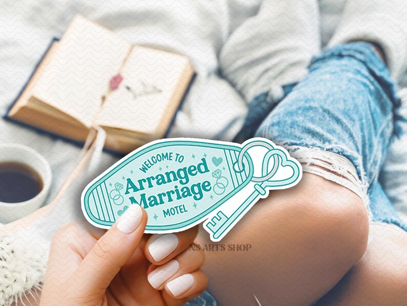 May include: A teal and white motel key tag with the words "WELCOME TO Arranged Marriage MOTEL" and a key design. The key tag is held in a hand with white nail polish. A book and a cup of coffee are in the background.