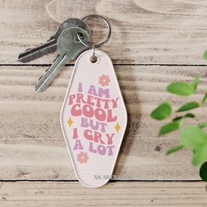 May include: A retro-style keychain with the text "I AM PRETTY COOL BUT I CRY A LOT" in pink, orange, and purple. The keychain has a light pink background with flower and diamond accents. Silver keys are attached to the keychain.