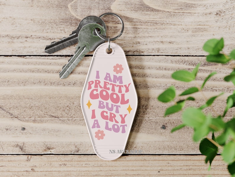 May include: A retro-style keychain with the text "I AM PRETTY COOL BUT I CRY A LOT" in pink, orange, and purple. The keychain has a light pink background with flower and diamond accents. Silver keys are attached to the keychain.