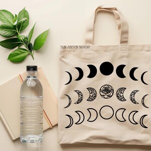 May include: A beige tote bag with black moon phases and a floral design. The bag has the text "NS ARTS SHOP" printed on the front.