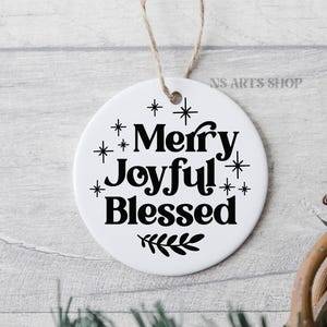 以下が含まれることがあります： 黒い文字で「Merry Joyful! Blessed」と書かれた白い丸いオーナメント。下部に小さな葉の枝があり、文字の周りに黒い星が散りばめられています。