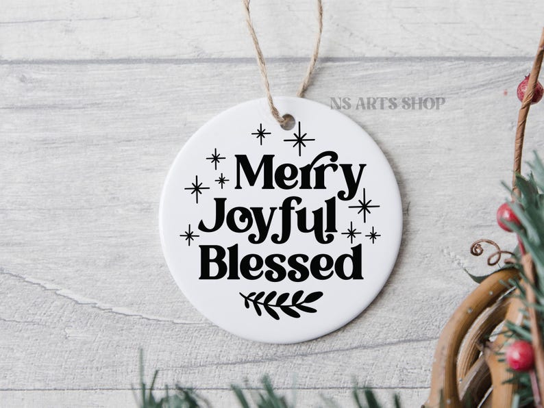 以下が含まれることがあります： 黒い文字で「Merry Joyful! Blessed」と書かれた白い丸いオーナメント。下部に小さな葉の枝があり、文字の周りに黒い星が散りばめられています。