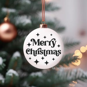 以下が含まれることがあります： 白いクリスマスオーナメントに黒い文字で「メリークリスマス」と書いてあり、黒い星形のアクセントが付けられています。