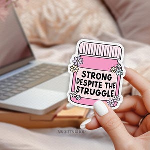 Peut inclure: Autocollant rose avec un pot et des fleurs. Le pot porte l'inscription "Strong despite the struggle".
