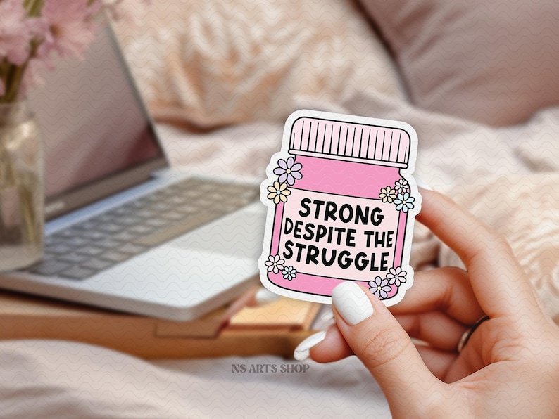 Peut inclure: Autocollant rose avec un pot et des fleurs. Le pot porte l'inscription "Strong despite the struggle".
