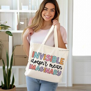 Peut inclure: Un sac fourre-tout blanc avec le texte "INVISIBLE doesn't mean IMAGINARY" en couleurs arc-en-ciel. Le sac est tenu par une personne portant une chemise blanche et un jean bleu.