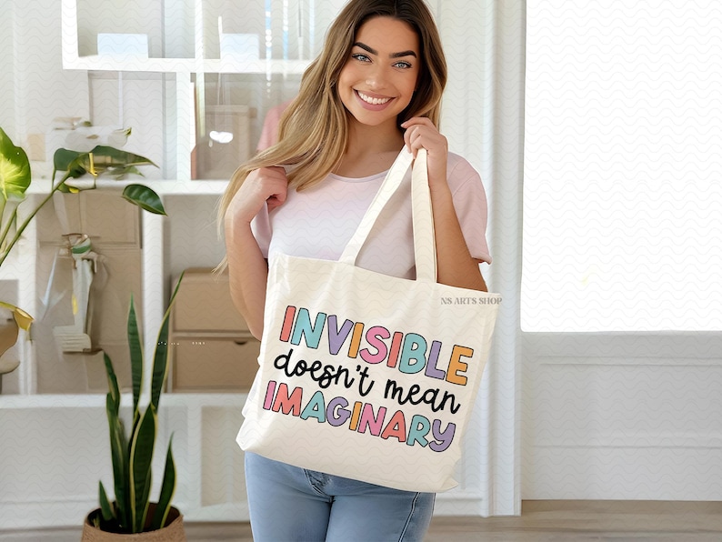 Peut inclure: Un sac fourre-tout blanc avec le texte "INVISIBLE doesn't mean IMAGINARY" en couleurs arc-en-ciel. Le sac est tenu par une personne portant une chemise blanche et un jean bleu.