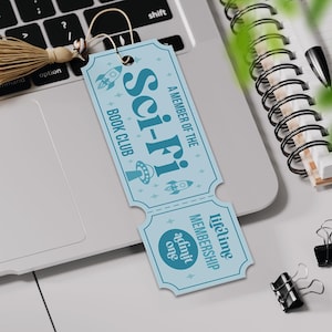 May include: A light blue bookmark with the words "Sci-Fi Book Club" and rocket ship and UFO illustrations. The bookmark has a tassel and the text "Lifetime Membership admit one". The bookmark is on a laptop.