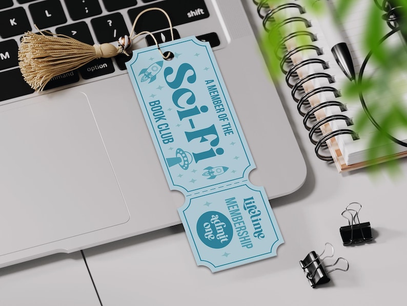 May include: A light blue bookmark with the words "Sci-Fi Book Club" and rocket ship and UFO illustrations. The bookmark has a tassel and the text "Lifetime Membership admit one". The bookmark is on a laptop.