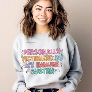 Peut inclure: Un sweat-shirt gris clair avec le texte "PERSONALLY VICTIMIZED BY MY IMMUNE SYSTEM" en lettres color&eacute;es avec des fleurs et des &eacute;toiles autour du texte.