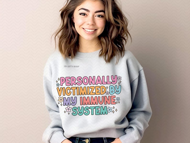 Peut inclure: Un sweat-shirt gris clair avec le texte "PERSONALLY VICTIMIZED BY MY IMMUNE SYSTEM" en lettres color&eacute;es avec des fleurs et des &eacute;toiles autour du texte.