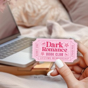 Puede incluir: Un boleto rosa con el texto "Dark Romance Book Club Lifetime Membership" y gr&aacute;ficos florales y de calaveras. El boleto se sostiene en una mano, con un port&aacute;til y flores en el fondo.