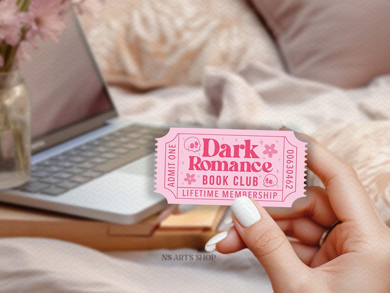Puede incluir: Un boleto rosa con el texto "Dark Romance Book Club Lifetime Membership" y gr&aacute;ficos florales y de calaveras. El boleto se sostiene en una mano, con un port&aacute;til y flores en el fondo.