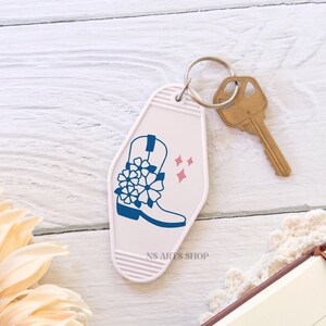 May include: A white motel-style keychain with a blue cowboy boot design and pink star accents. A gold-colored key is attached to the keychain. The text "NS ARTS SHOP" is printed on the keychain.