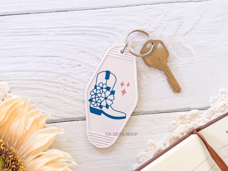 May include: A white motel-style keychain with a blue cowboy boot design and pink star accents. A gold-colored key is attached to the keychain. The text "NS ARTS SHOP" is printed on the keychain.