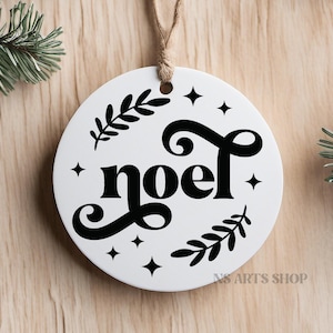 以下が含まれることがあります： 黒い文字で「noel」とスタイリッシュなフォントで書かれた白い丸いクリスマスオーナメント。オーナメントは小さな星と緑の葉で飾られています。