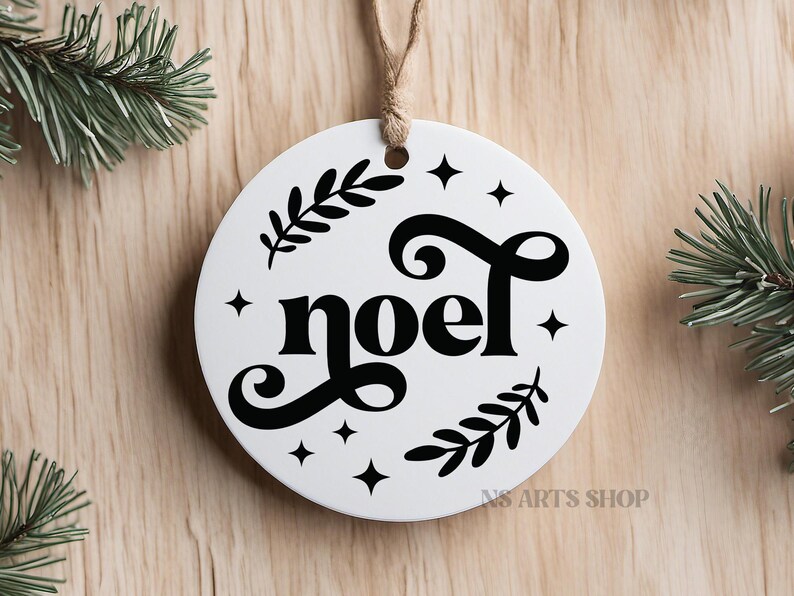 以下が含まれることがあります： 黒い文字で「noel」とスタイリッシュなフォントで書かれた白い丸いクリスマスオーナメント。オーナメントは小さな星と緑の葉で飾られています。