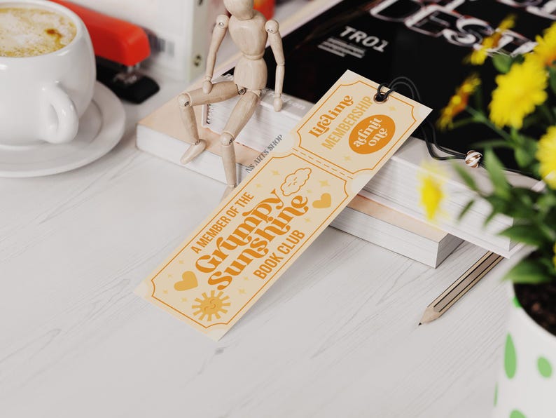 Op de afbeelding: Een oranje en bruine bladwijzer met de tekst "A Member of the Grumpy Sunshine Book Club" en "Lifetime Membership Admit One". De bladwijzer ligt op een stapel boeken, met een kopje koffie en bloemen op de achtergrond.