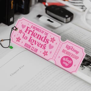 Op de afbeelding: Een roze bladwijzer met de tekst "A Member of the Friends to Lovers Book Club" en "Lifetime Membership admit one". De bladwijzer is versierd met hartjes en bloemen en ligt in een open boek.