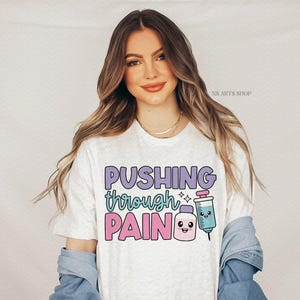 Peut inclure: T-shirt blanc avec le texte "Pushing through pain" en lettres violettes, roses et bleues. Une seringue de dessin anim&eacute; avec un visage souriant est &agrave; c&ocirc;t&eacute; du texte.