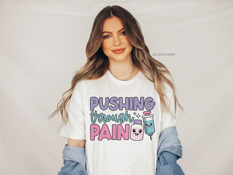 Peut inclure: T-shirt blanc avec le texte "Pushing through pain" en lettres violettes, roses et bleues. Une seringue de dessin anim&eacute; avec un visage souriant est &agrave; c&ocirc;t&eacute; du texte.