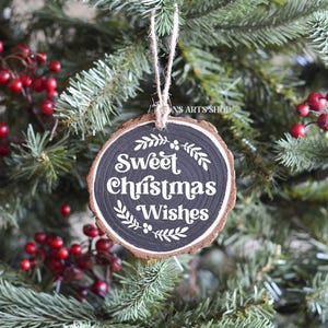 以下が含まれることがあります： 黒い背景に白い文字で「Sweet Christmas Wishes」と書かれた丸い木製のクリスマスオーナメント。オーナメントは赤い実のついたクリスマスツリーに飾られています。