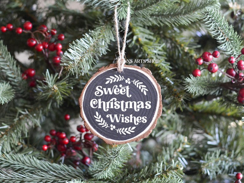 以下が含まれることがあります： 黒い背景に白い文字で「Sweet Christmas Wishes」と書かれた丸い木製のクリスマスオーナメント。オーナメントは赤い実のついたクリスマスツリーに飾られています。