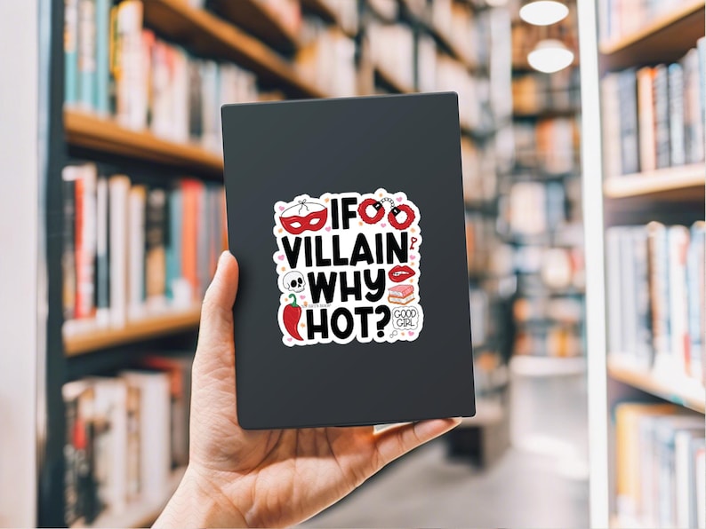 Puede incluir: Adhesivo negro con el texto "IF VILLAIN WHY HOT?" e ilustraciones variadas, incluyendo una m&aacute;scara, esposas, una calavera, un chile y un par de labios. El adhesivo se sostiene en una mano sobre un fondo borroso de estanter&iacute;as.